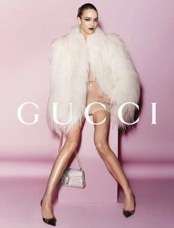 Fashion shot: A model in a cream fur coat poses against a pink backdrop, holding a glittering purse with a large logo. Gucci logo displayed.