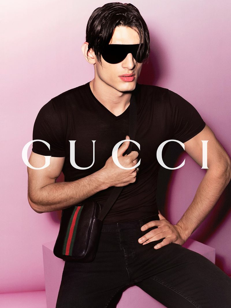 A man poses with a black leather satchel bag in a fashion shot on a light pink backdrop. He wears sunglasses and all-black attire.
