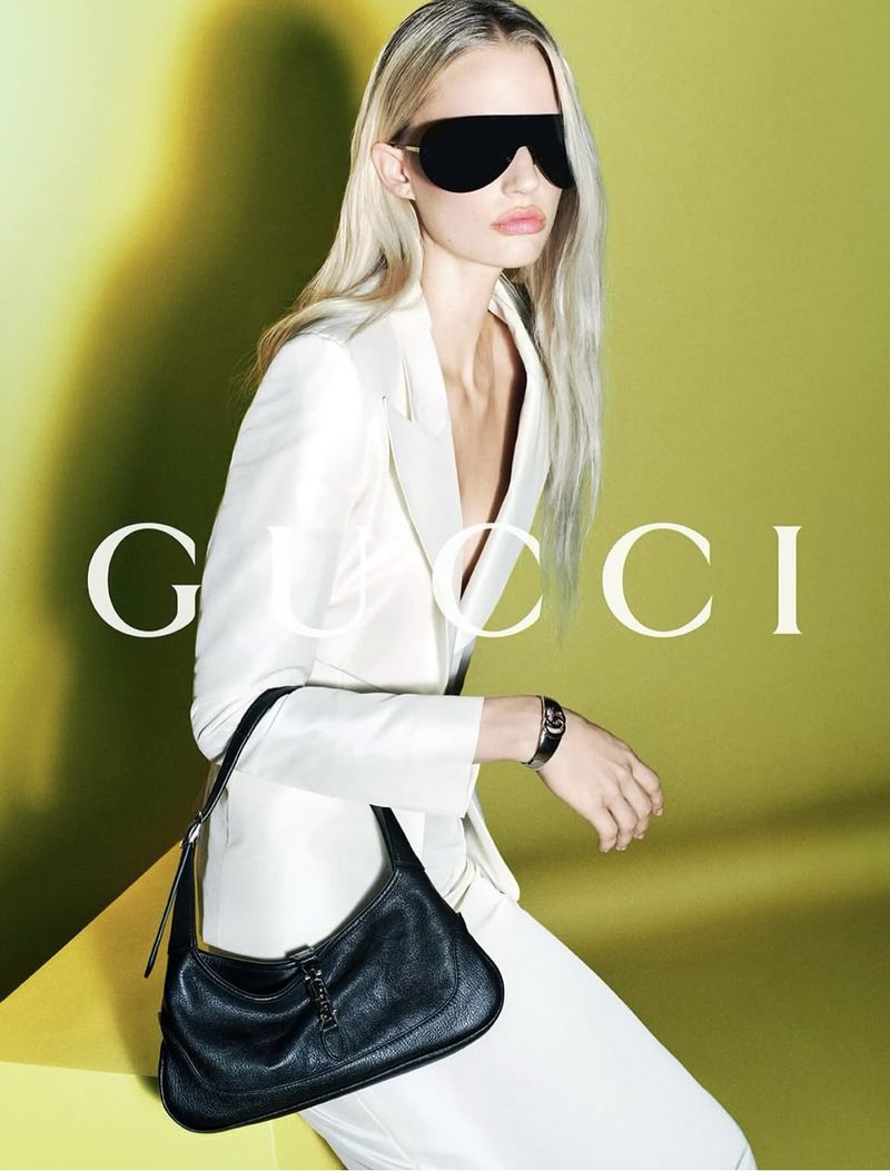 A model wearing a white jacket and sunglasses poses in front of a lime green and yellow background, holding a black Gucci purse.