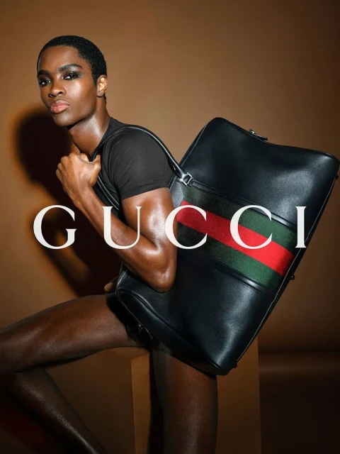 Dark-skinned male model poses with a large black Gucci bag featuring red and green stripes against a warm beige background.