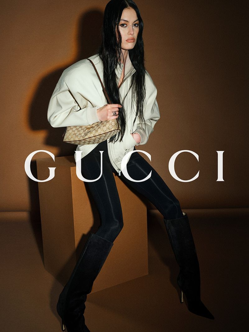 Fashion shoot featuring a model in a cream jacket, black leggings, and high boots, holding a Gucci bag in a studio setting.