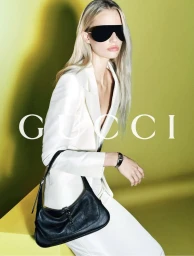 Model wearing dark shield sunglasses and a white tailored suit holding a black leather shoulder bag against a chartreuse background.