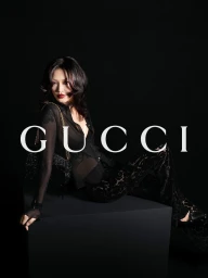 Model in a sheer black top and patterned trousers reclining on a black block with the white Gucci logo overlaid.