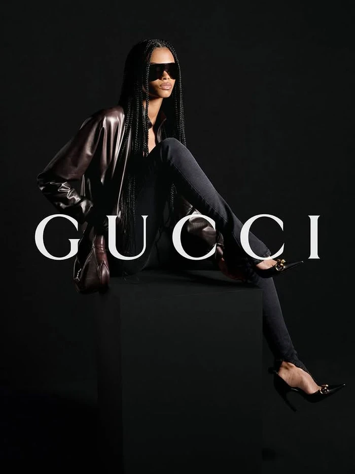 A fashion model wearing black sunglasses, a brown leather jacket, and stilettos sits on a black cube against a black background.