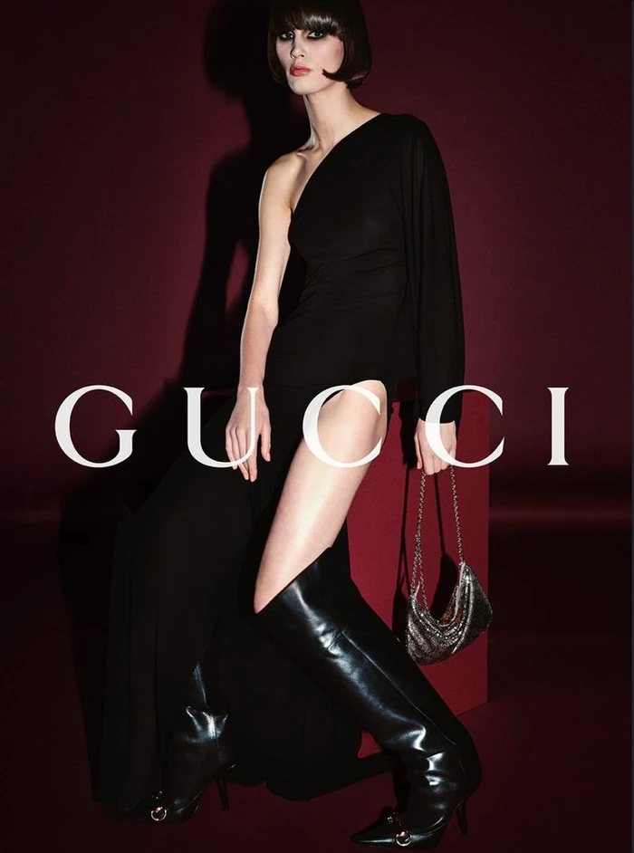 Model wearing a black one-shoulder high-slit gown and tall leather boots against a deep burgundy backdrop for Gucci.