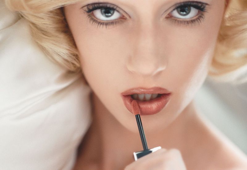 Close-up of a blond woman applying lip gloss with a wand applicator, soft light, portrait orientation, beauty shot, pale skin.