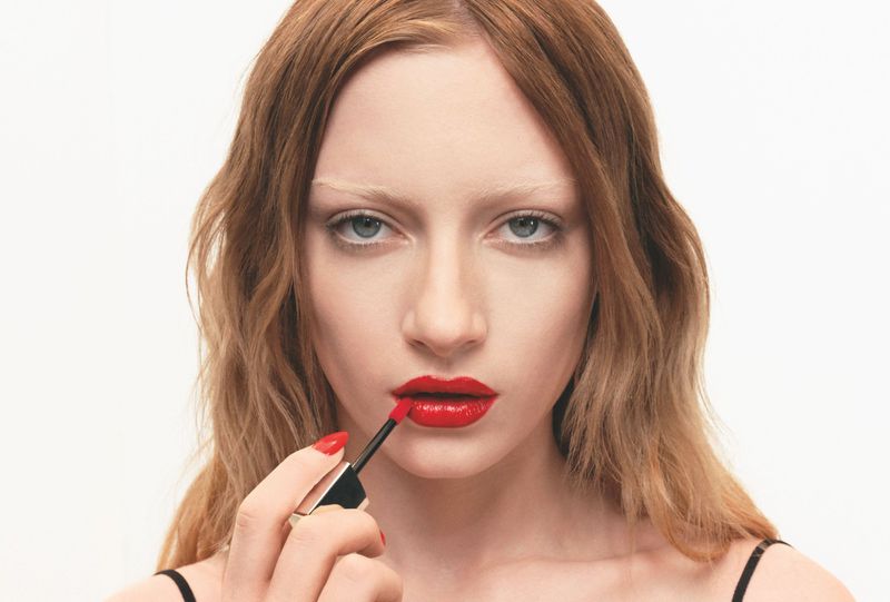 Close-up shot of an adult female model with red lipstick, holding a lip gloss applicator. Red nail polish matches the lipstick.