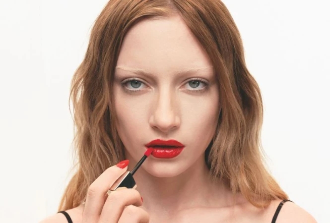 Close-up shot of an adult female model with red lipstick, holding a lip gloss applicator. Red nail polish matches the lipstick.