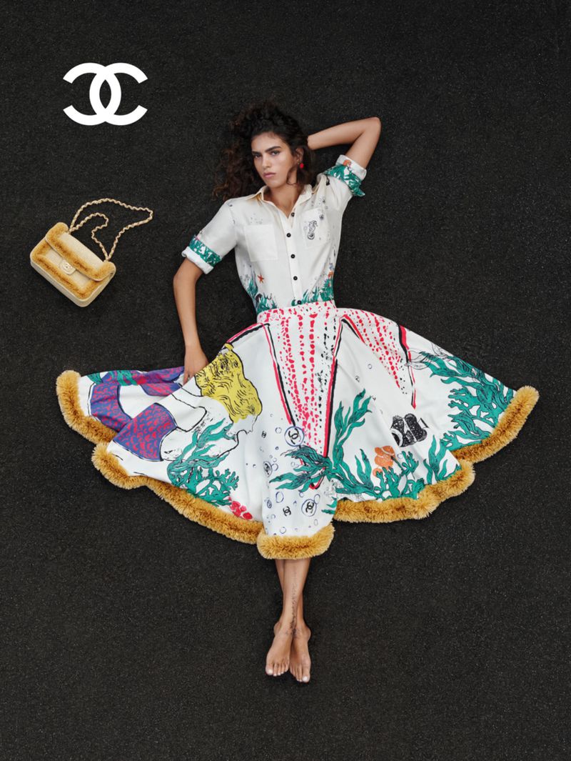 Woman lies on a black surface wearing a white blouse and illustrated skirt with a fur purse and the Chanel logo in the upper left.