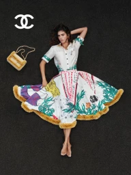 Model lying on dark textured pavement wearing an illustrated printed dress with fur trim beside a yellow shearling handbag.