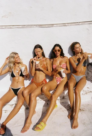 Four women in swimsuits pose outside, drinking beverages and sitting together against a white wall in sunny, natural lighting.