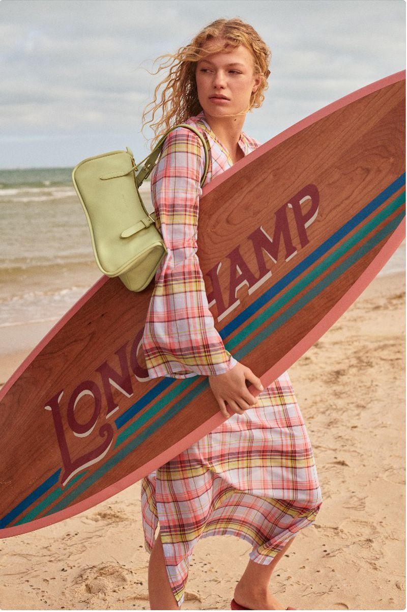 An adult woman with curly blonde hair stands on a beach holding a surfboard and wearing a plaid dress with a green purse slung over her shoulder.