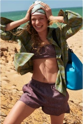 Model Liva Borre smiling on a sunny beach wearing a green plaid shirt, purple shorts set, headscarf, and blue shoulder bag.