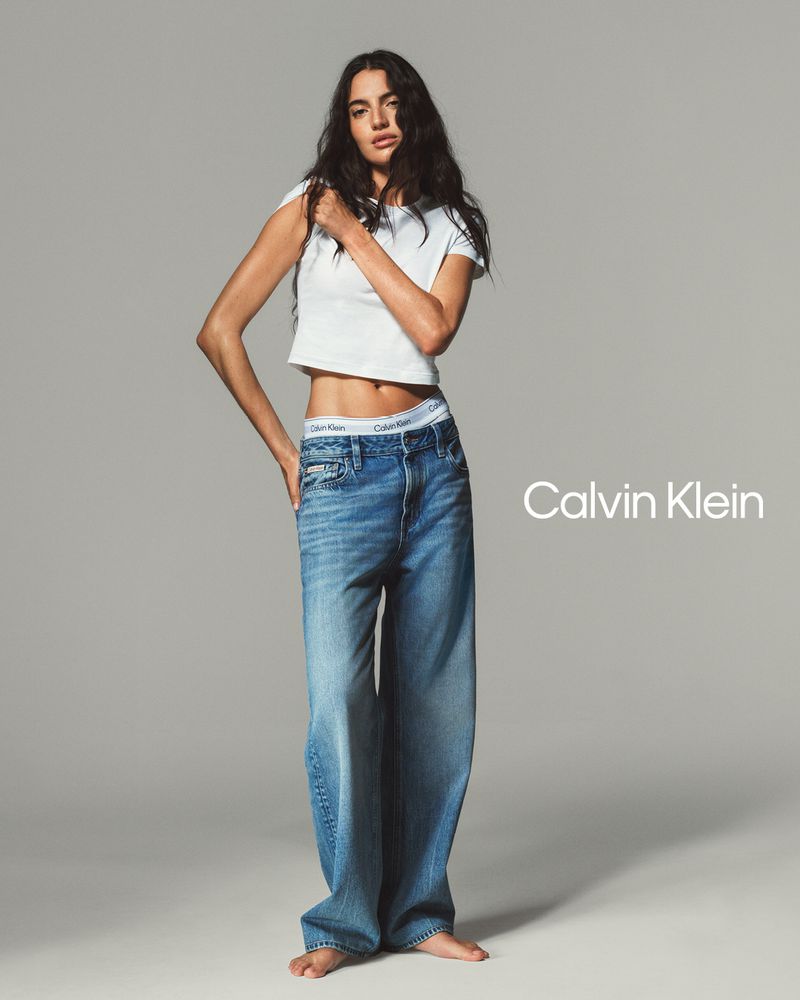 Brunette woman poses in a white t-shirt and wide leg jeans in a studio shot with a grey background and visible Calvin Klein logo.