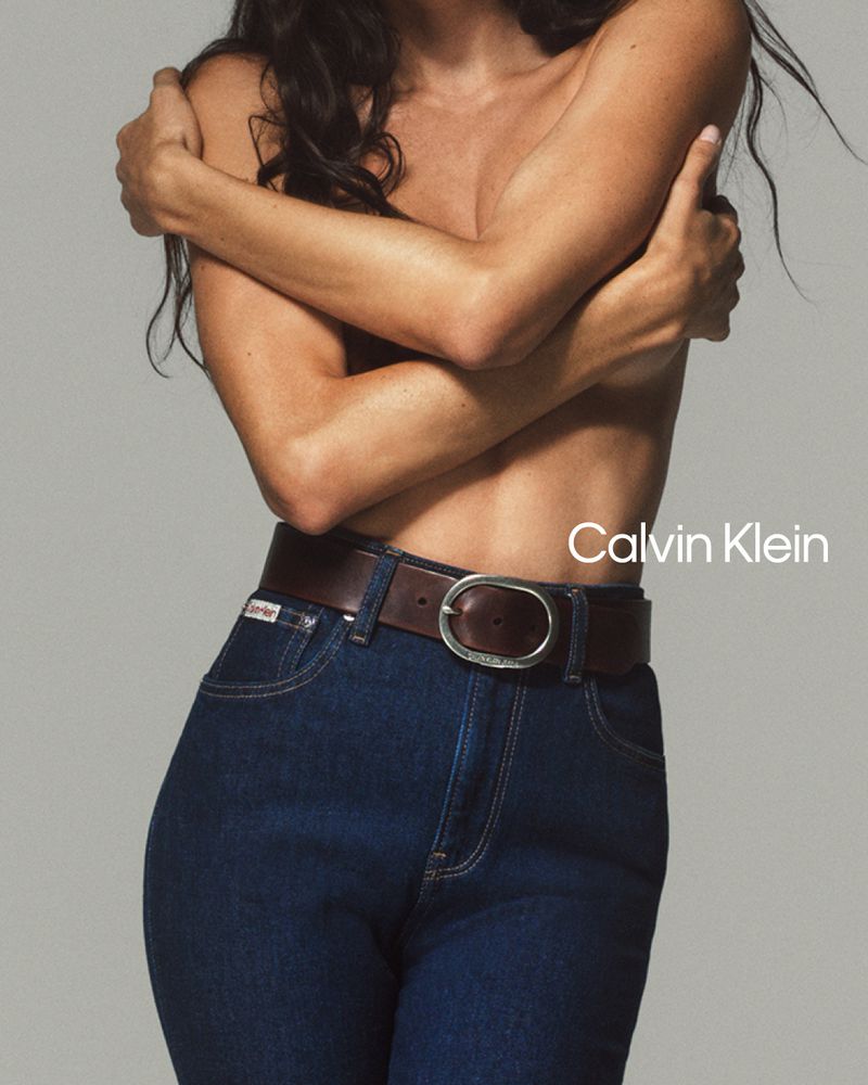 A young woman with long hair wears dark blue jeans and a brown belt, posing against a neutral grey background. The Calvin Klein logo is visible.