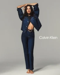 Model posing barefoot in a dark blue denim jacket and matching jeans with an oval buckle belt against a grey studio backdrop.