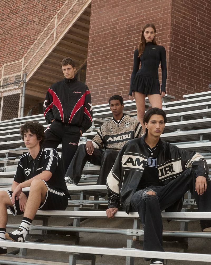 Five fashion models pose on bleachers in stylish jackets, shirts, and skirts, showcasing apparel with brand logo in front of a brick wall.