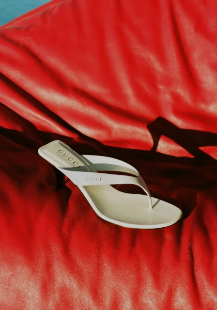 Elevated shot of a white high-heeled Gucci sandal atop a draped red fabric, capturing shadows and brand visibility in a studio setting.