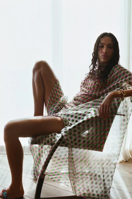 Adult female model poses in chair wearing printed dress and slides in an indoor setting with natural window light, fashion editorial.