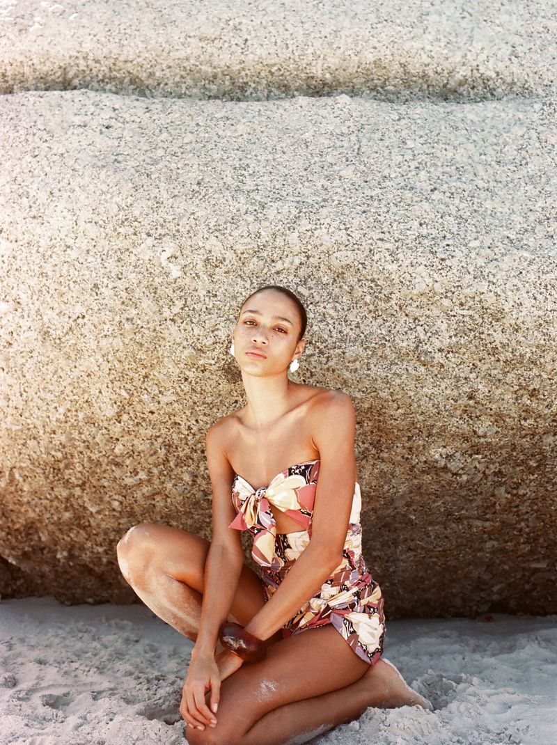 Woman in floral strapless romper sits on a sandy beach in front of a large rock, wearing earrings and bracelet in outdoor shot.