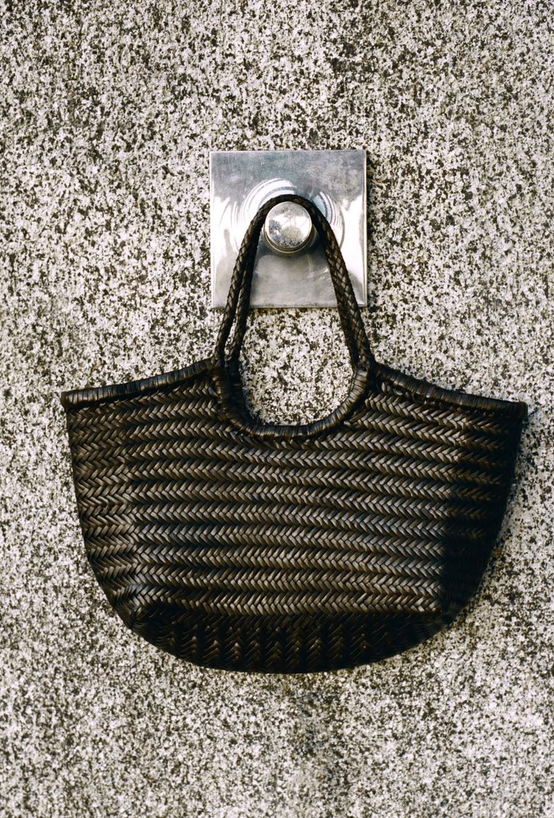 Woven straw bag hangs from a metal wall-mounted hook against a gray speckled wall in an eye-level, medium studio shot. Neutral lighting.