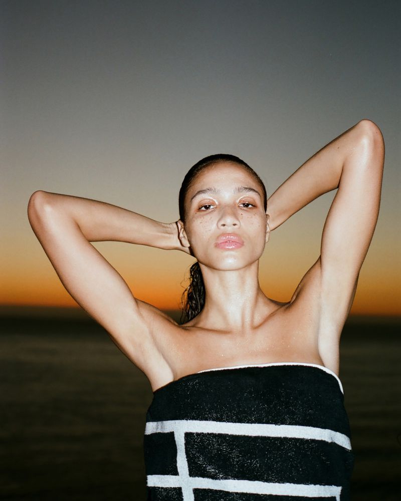 Portrait of a woman with dark hair wrapped in a patterned towel, arms above head, standing outdoors against an orange sunset background.