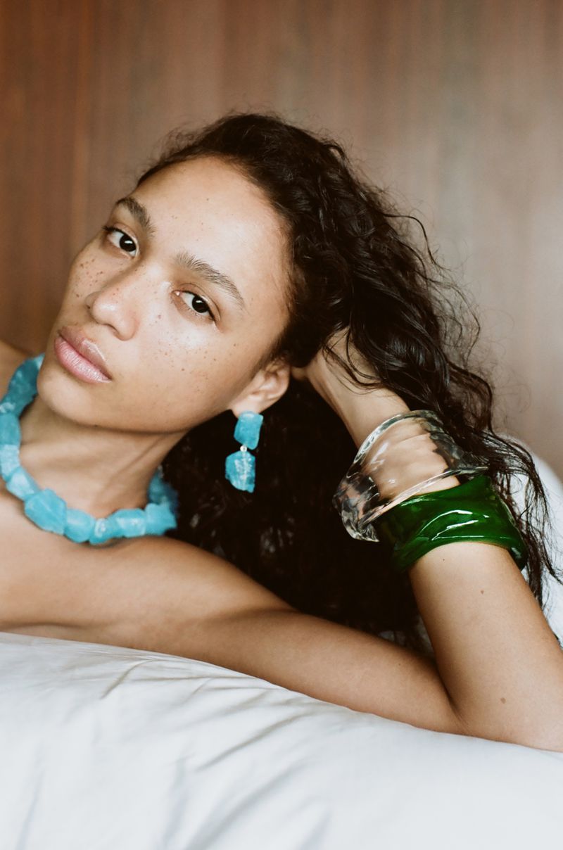 Portrait of a woman with dark curly hair wearing a blue stone necklace, matching earrings, and green bangle bracelets lying on a white surface.