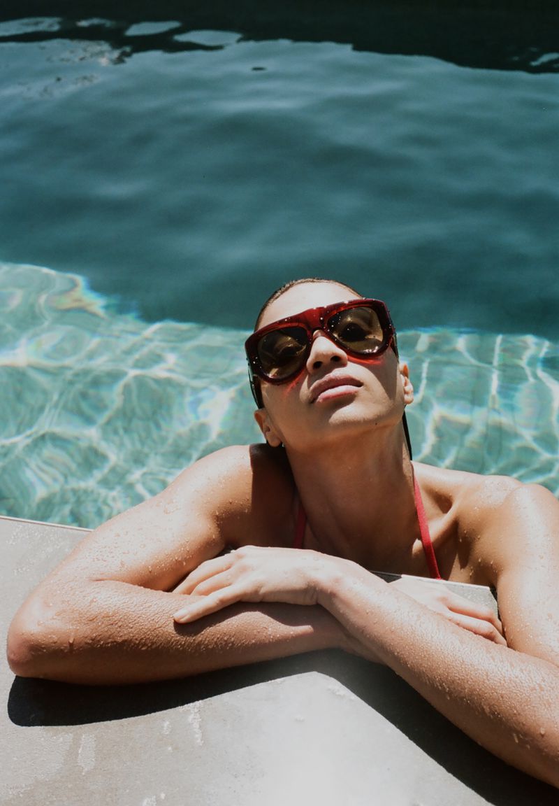 Woman wearing sunglasses and a swimsuit in a pool on a sunny day. She is resting her arms on the edge of the pool.