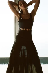 Model Mauria Caetano poses in a black cutout one-piece swimsuit and sheer black maxi skirt against a bright pool window.