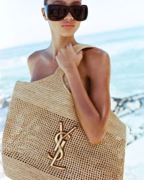 Model wearing oversized black sunglasses carrying a woven raffia tote bag with gold logo hardware at the beach.