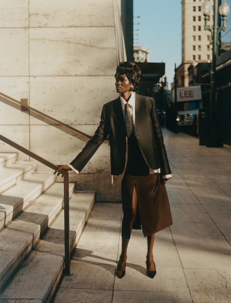 Elegant black woman poses in a suit and brown skirt on city steps. The image features a modern, urban setting with natural lighting.