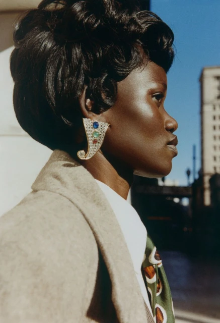 Profile view of an adult woman with short dark hair and large decorative earrings wearing a patterned tie and tan blazer outdoors.