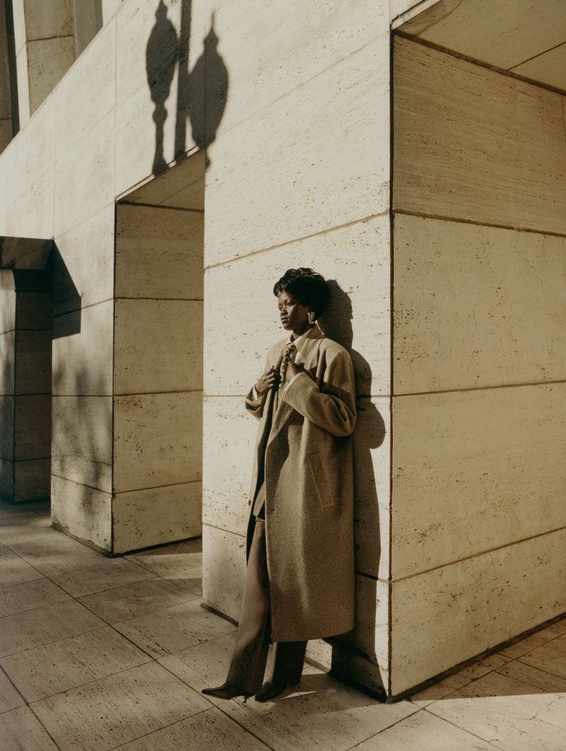 An African-American woman in a tan coat poses against a travertine building. The sunlight casts shadows of her and a lamppost.