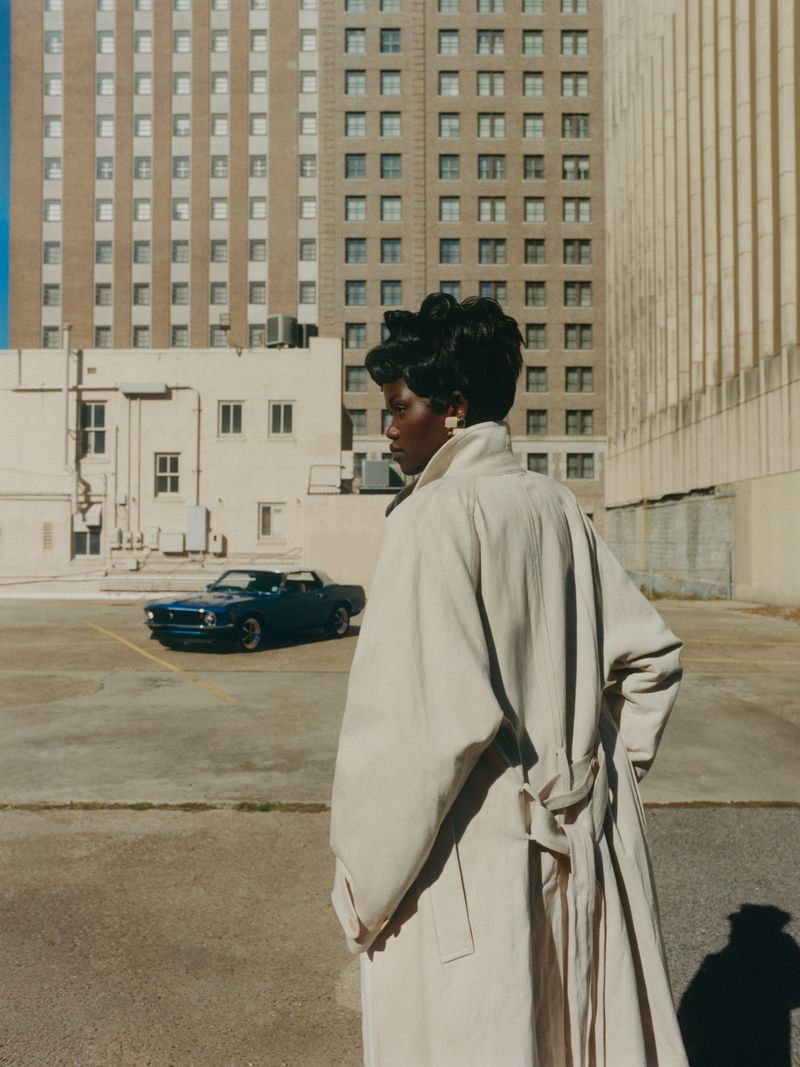 Woman in tan trench coat stands in outdoor parking area in front of blue vintage car and a tall building with many windows.