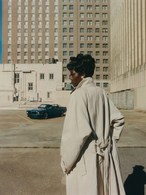 Woman in tan trench coat stands in outdoor parking area in front of blue vintage car and a tall building with many windows.