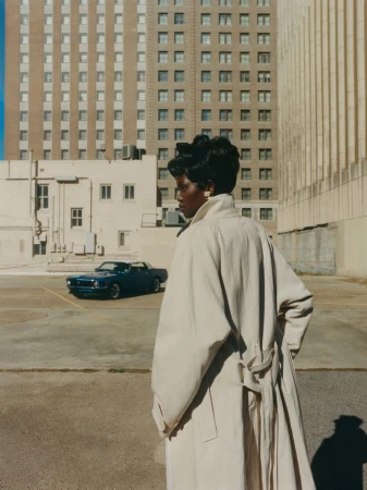 Woman in tan trench coat stands in outdoor parking area in front of blue vintage car and a tall building with many windows.