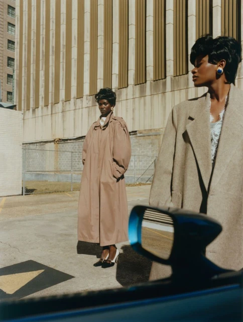 Two women in trench coats stand outside a building, one reflected in a car mirror. Urban fashion photography with natural lighting.
