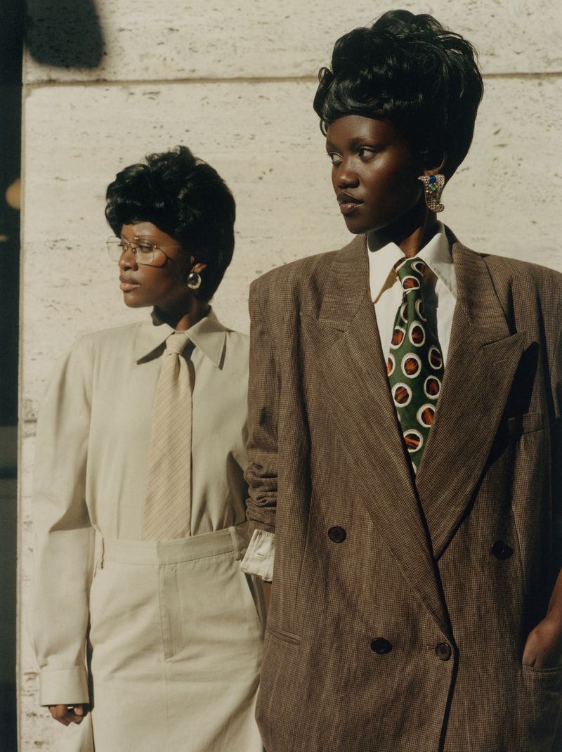 Two adult women pose in vintage suits against an off-white textured background with natural light, showcasing classic fashion apparel.