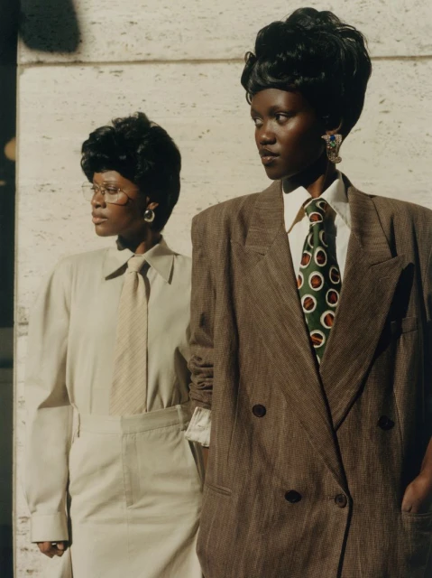 Two adult women pose in vintage suits against an off-white textured background with natural light, showcasing classic fashion apparel.