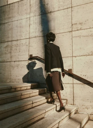 Adult female in black jacket and brown skirt ascends a concrete staircase outdoors in warm, natural light casting strong shadows.