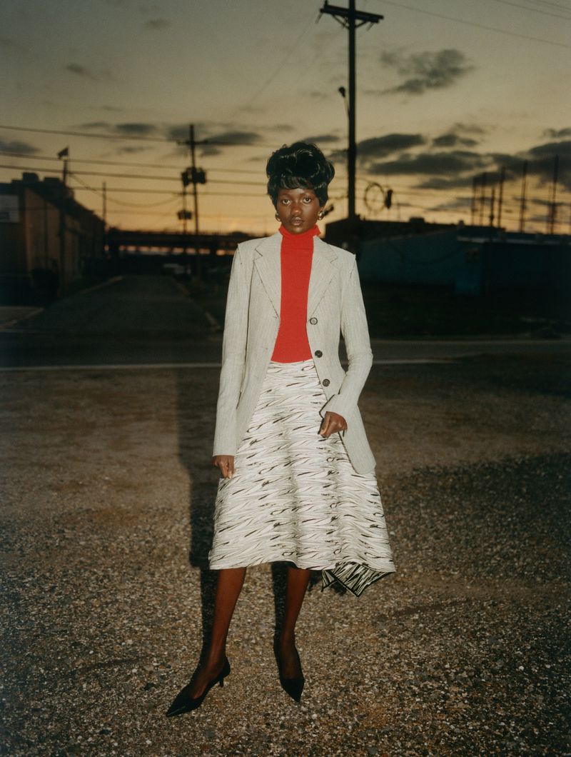 Woman in coat and skirt posing outdoors with buildings and power lines visible against a sunset. Fashion editorial photography.