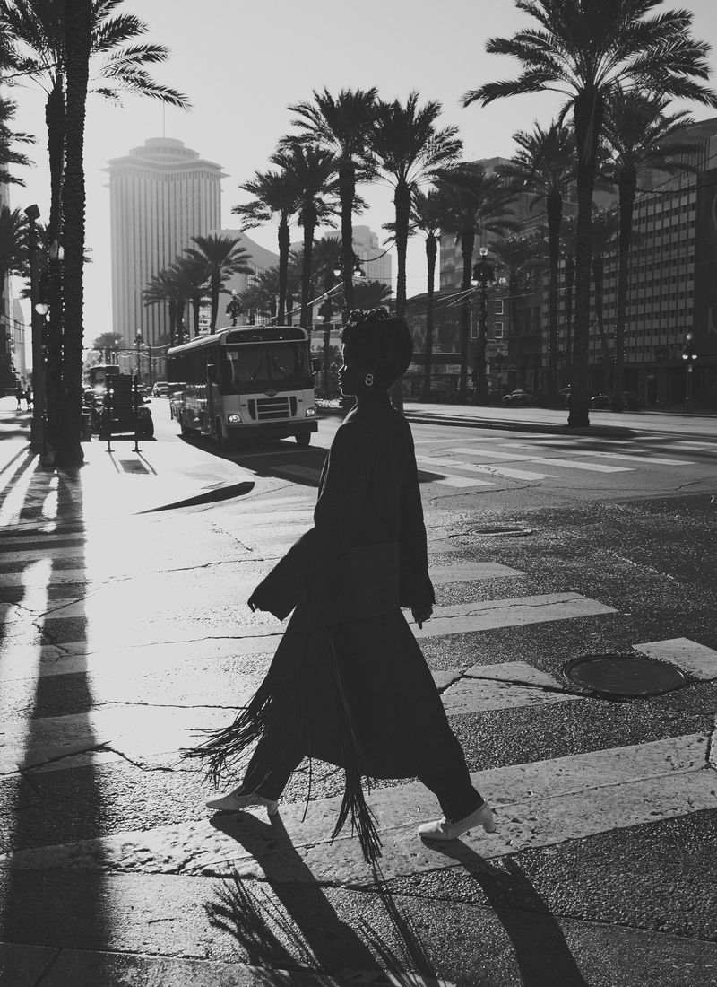 Monochrome image of a woman crossing a street in a city with palm trees and buildings, captured with natural light and shadows.