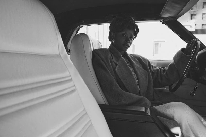 Elegant Black woman driving a classic car with white interior; city buildings visible in the background, monochrome color palette.
