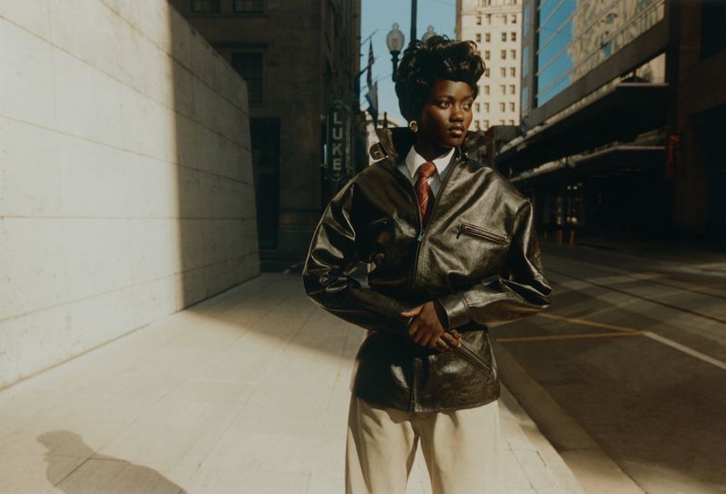 A stylish African-American woman in a leather jacket poses on a city street, with skyscrapers and buildings in the background, bathed in sunlight.
