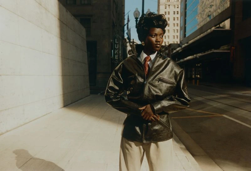 A stylish African-American woman in a leather jacket poses on a city street, with skyscrapers and buildings in the background, bathed in sunlight.