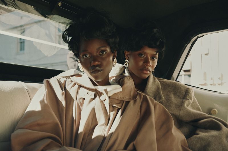 Two stylish Black women in overcoats are inside a car, looking directly at the camera with serious expressions. Fashion editorial aesthetic.
