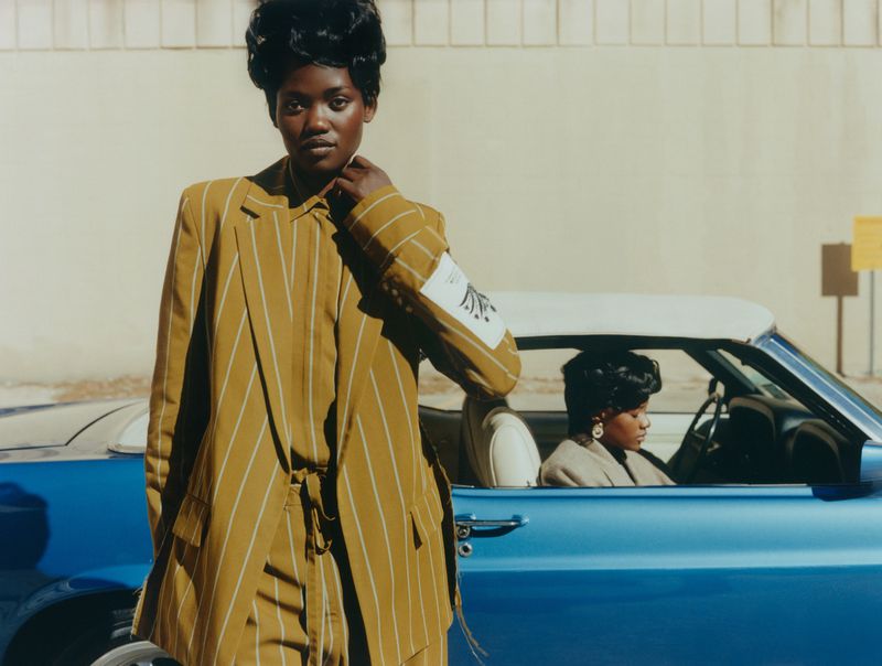 Two African American women pose next to a blue convertible. One woman in a striped suit stands outside the car, the other drives.