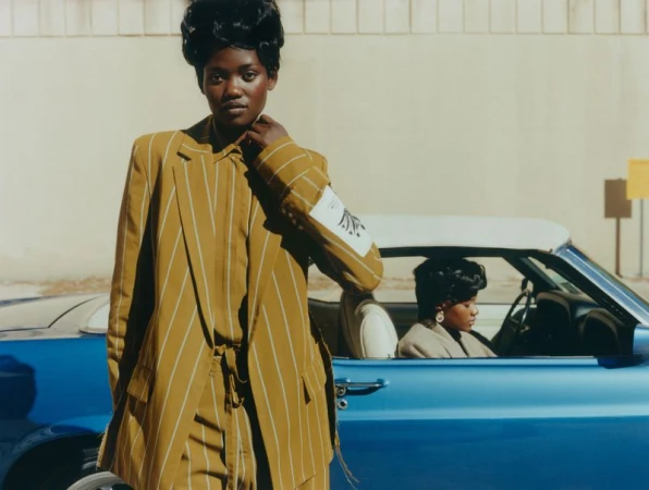 Two African American women pose next to a blue convertible. One woman in a striped suit stands outside the car, the other drives.