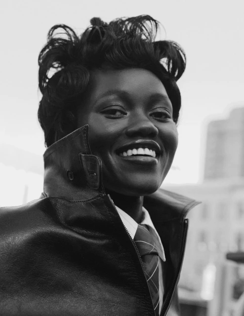 Close-up black and white portrait of smiling African American adult woman with short curly hair, wearing a collared shirt, tie, and leather jacket.