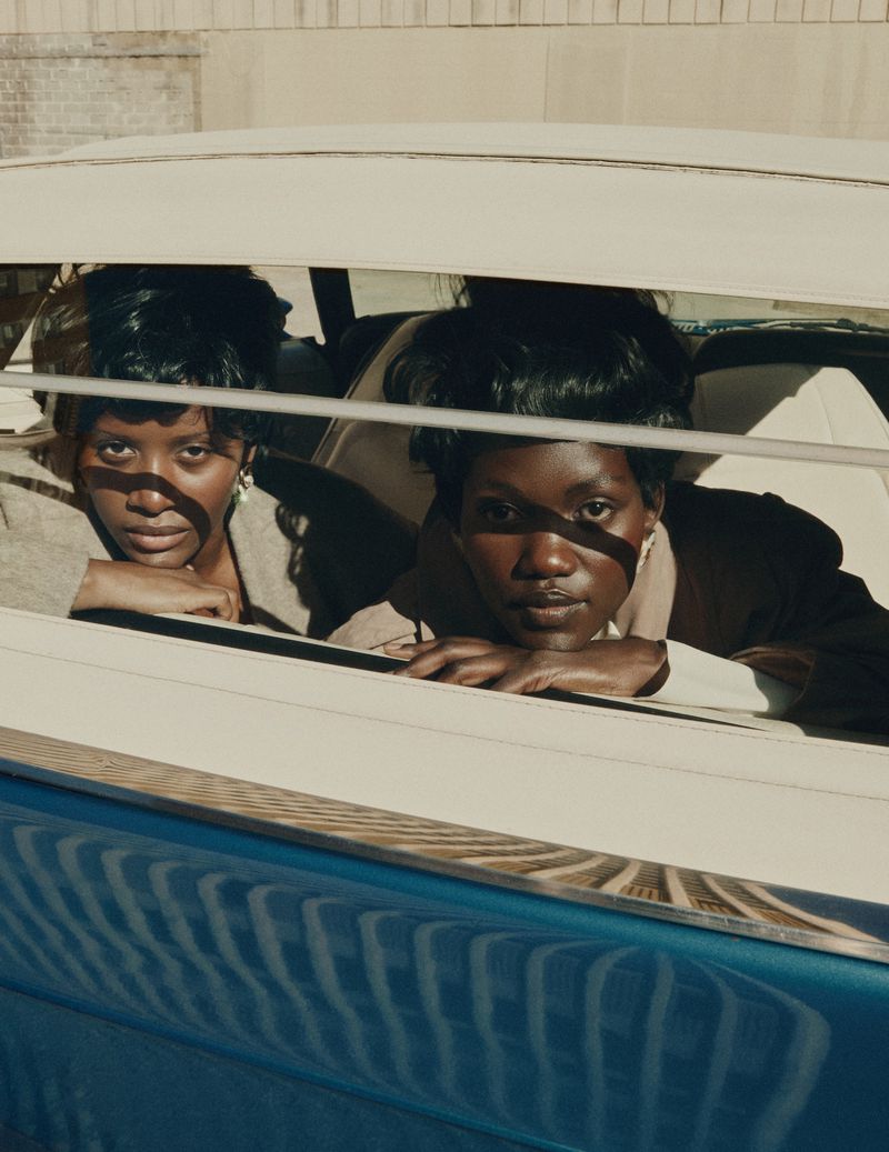 Two African American women in a blue car, wearing blazers, lean against the car frame, looking directly at the camera with neutral expressions.